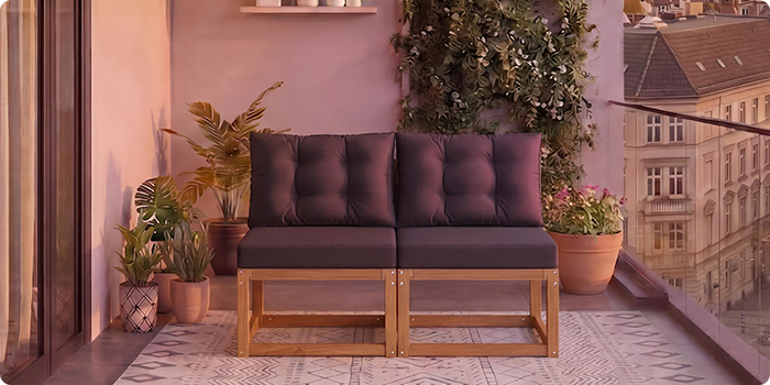 Divvietīgs modulārs koka dīvāns ar tumši pelēkiem spilveniem uz balkona, ko ieskauj podu augi un fona pilsētas skats